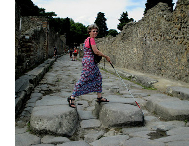 Photo shows a narrow street made of large flat stones, with a sidewalk about a foot high also made of large stones. Between the corners of the street is a row of 3 stepping stones, each about a foot high, 2 feet deep and 3 feet wide, spaced about a foot apart.  Dona is walking along the top of them, using a white cane and smiling at us.